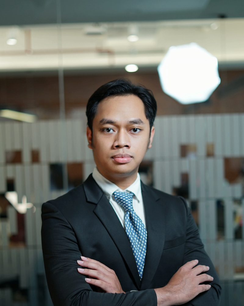 Portrait of Rafii Priadji in a dark suit and blue patterned tie, with arms crossed, in an office.